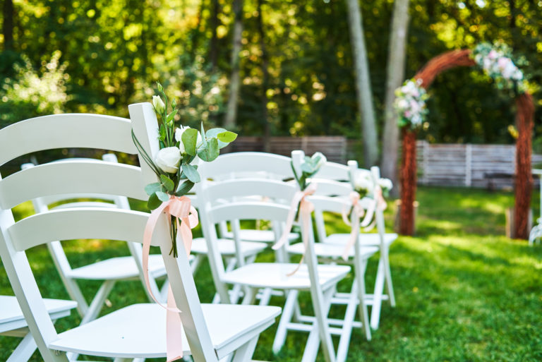 White fresh flowers with pink ribbon on wedding chair on each side of archway outroods; copy space. Wooden chairs for guests on green grass in the garden on wedding ceremony